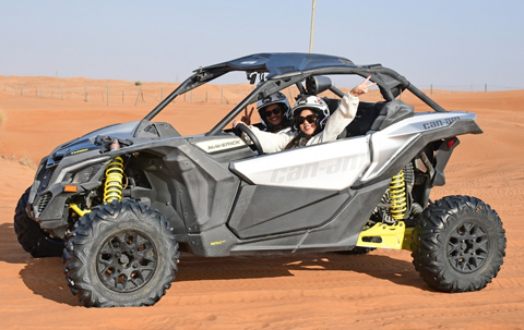 Smiling couple wearing helmets riding a Can-Am Maverick off-road buggy across the Dubai desert during an exciting dune buggy adventure.
