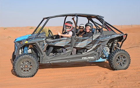 Three people riding a Polaris RZR off-road buggy across the Dubai desert during a guided dune adventure, wearing helmets and smiling at the camera.
